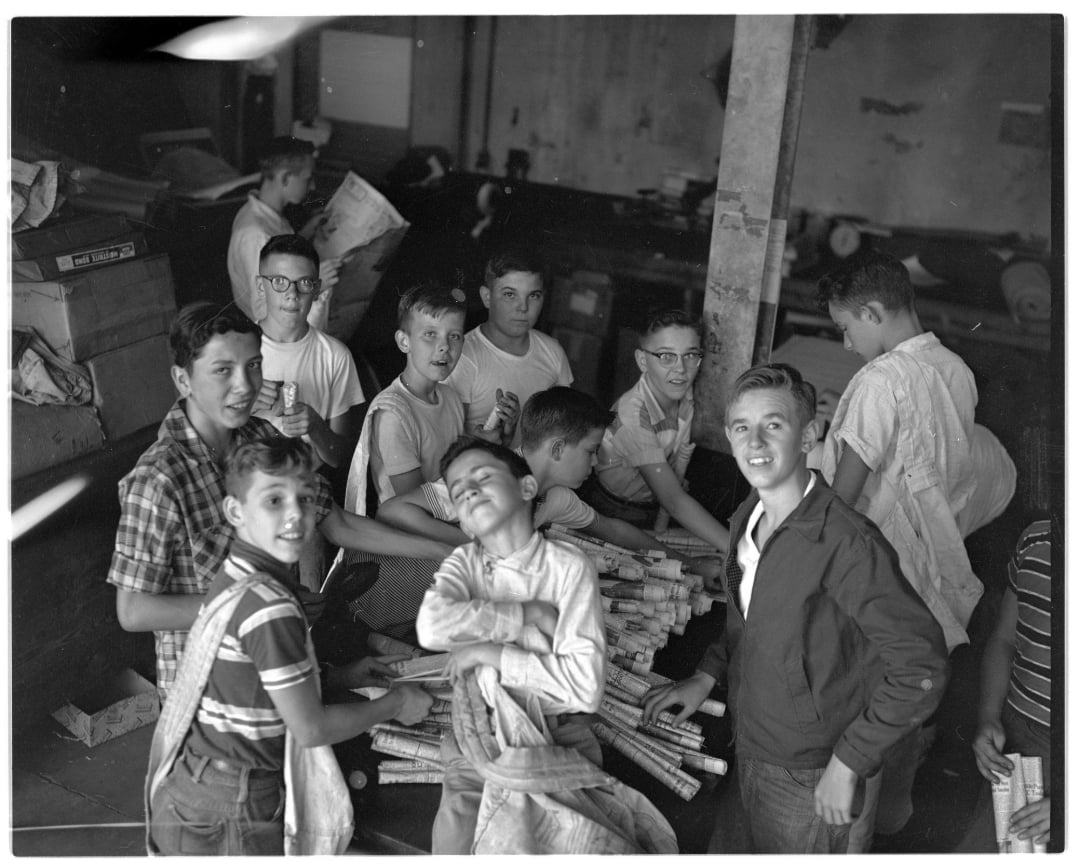 A group of boys, some smiling at the camera, gather indoors around bundles of rolled newspapers. The room appears cluttered with boxes and other items in the background.