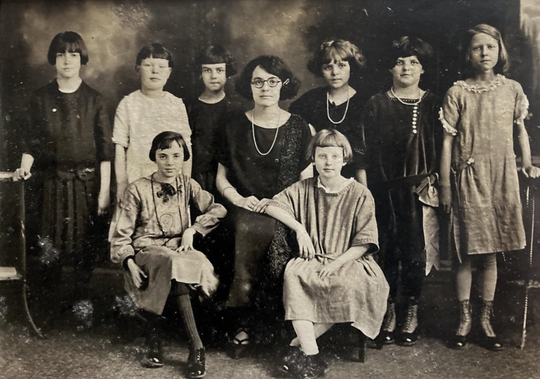 A vintage black and white photo showing nine girls and a woman sitting and standing in two rows, all dressed in early 20th-century clothing, posing formally and looking at the camera.