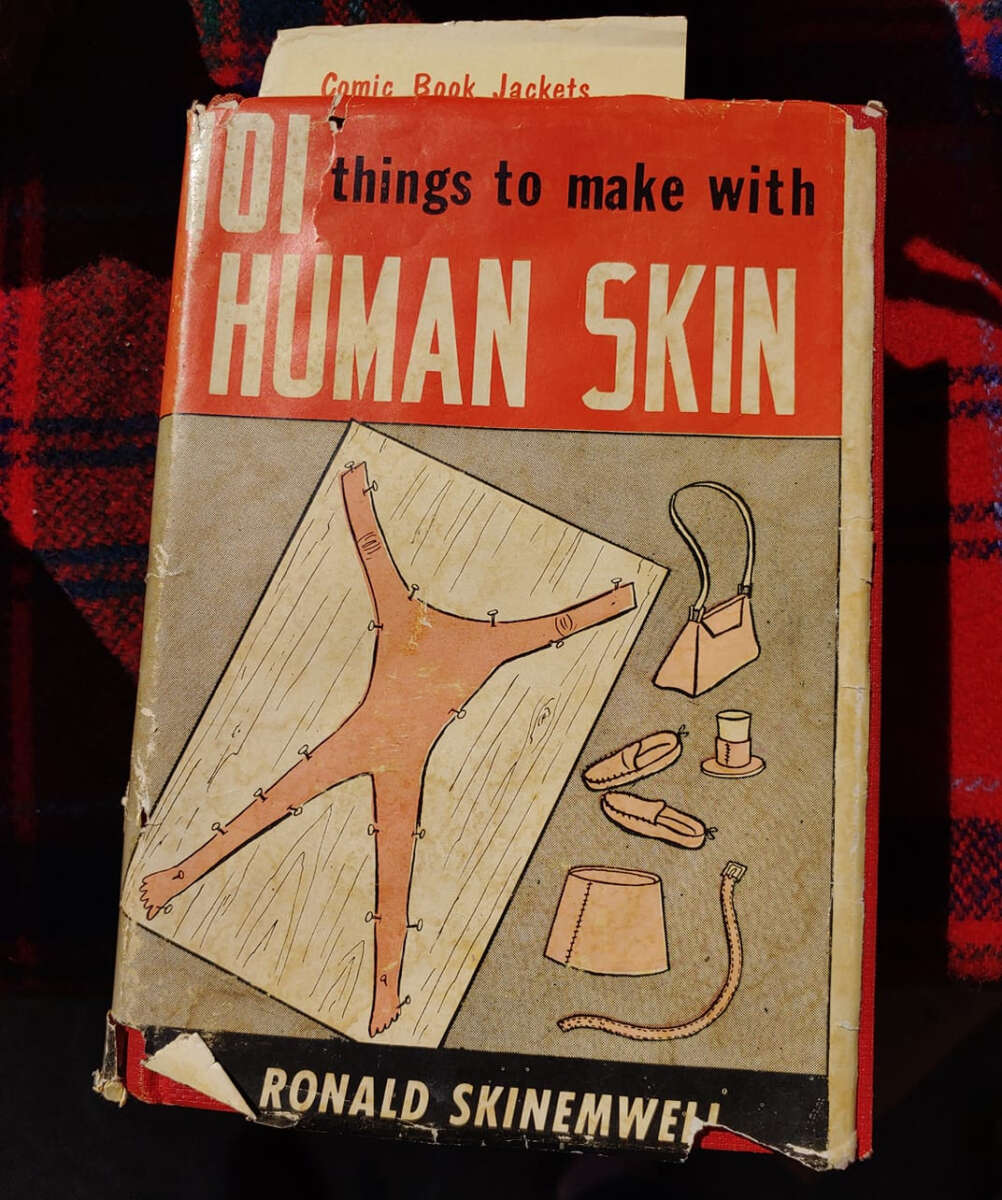 A worn book titled "101 things to make with HUMAN SKIN" by Ronald Skinewmeh. The cover shows a cartoon drawing of a stretched, stitched skin and various objects like a glove and lamp made from skin.