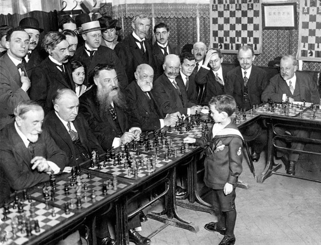 A young boy in a sailor suit plays simultaneous chess games against a group of seated older men, with a crowd of onlookers standing behind them in a room decorated with chessboards.