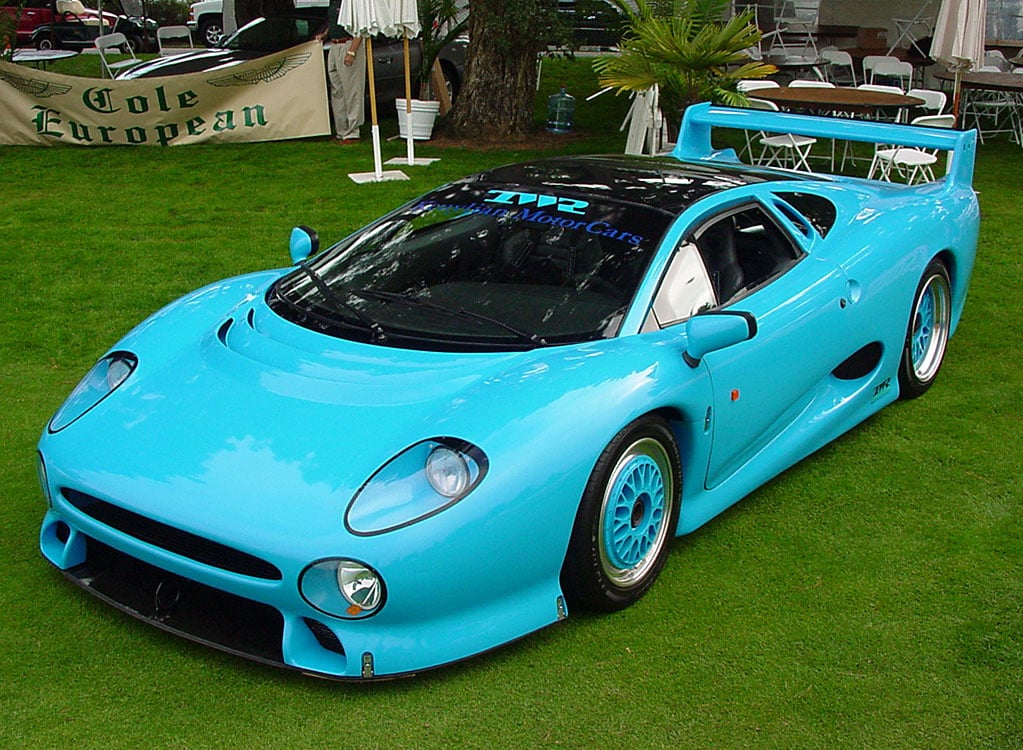 A bright blue Jaguar XJ220 sports car is displayed on a grassy lawn at an outdoor event, featuring a prominent rear wing, sleek aerodynamic design, and event tents in the background.