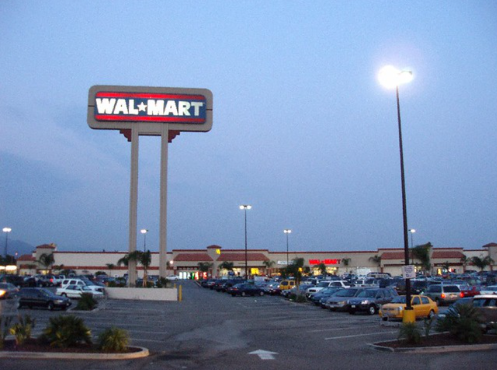 A large Walmart store at dusk with a tall illuminated sign, a well-lit parking lot filled with cars, and palm trees scattered around the lot.