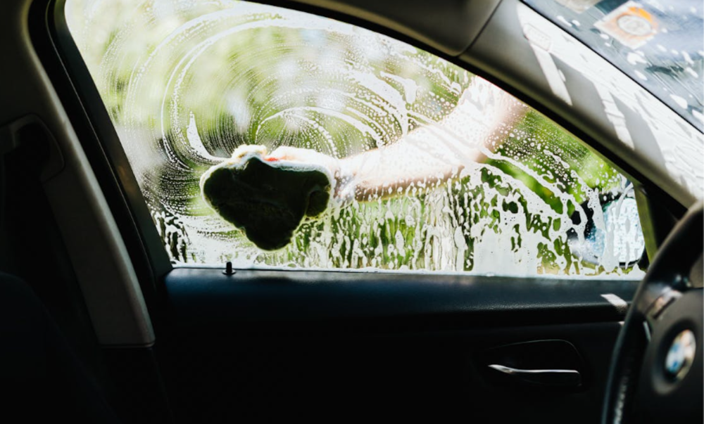A person cleans a car window from the outside using a green sponge, with soap and water creating swirls on the glass. The image is taken from inside the car, showing part of the door and interior.