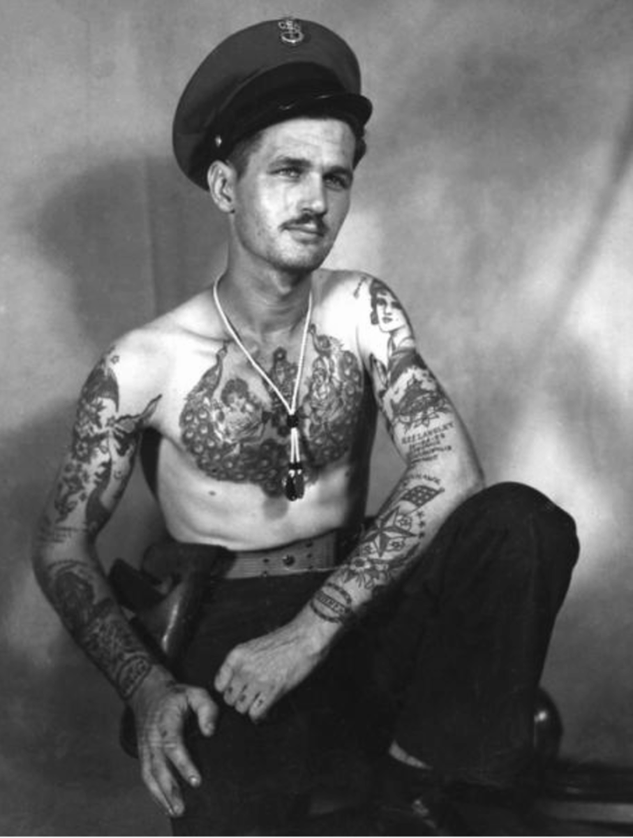 A black-and-white photo of a shirtless man with many tattoos, wearing a military-style cap and dark pants, posed sitting with one knee up, looking slightly off to the side.