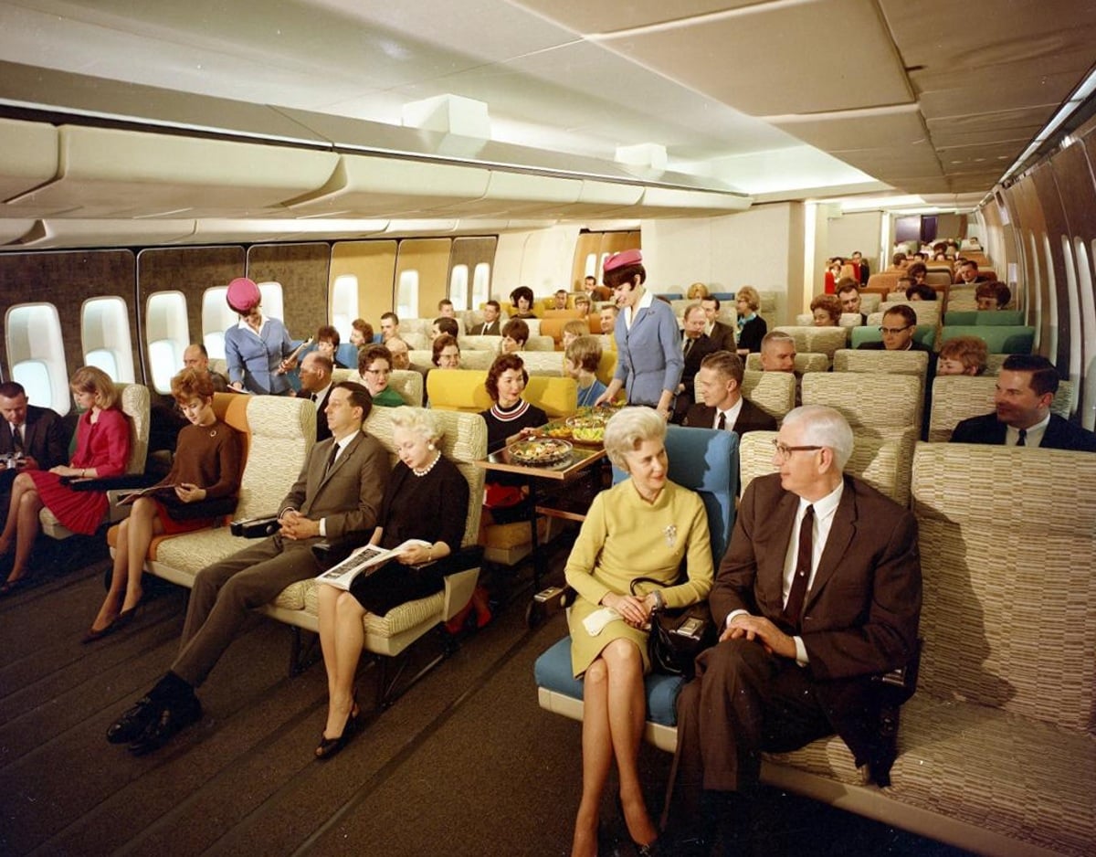 Passengers sit in a spacious, retro airplane cabin with colorful seats. Flight attendants in pink uniforms walk the aisles. Most passengers appear relaxed, reading or talking, with large windows and ample legroom visible.