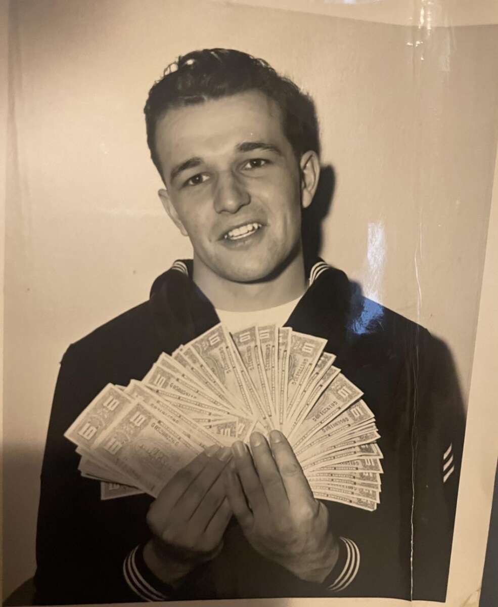 A young man in a dark uniform holds and fans out several paper bills while smiling at the camera. The photo is black and white.