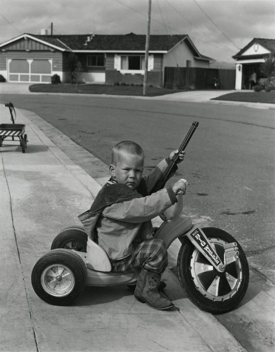 A young boy rides a Big Wheel tricycle on a suburban sidewalk, wearing boots and holding a toy rifle, with houses and a quiet street in the background.