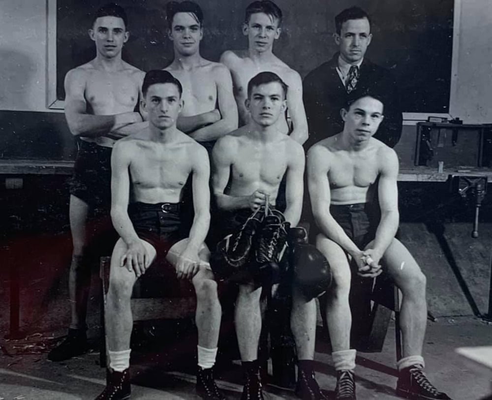 A black-and-white photo of six young men, five shirtless and wearing shorts and boots, sitting and standing in two rows. One man, dressed in a suit, stands at the back. The group appears to be a vintage sports team.