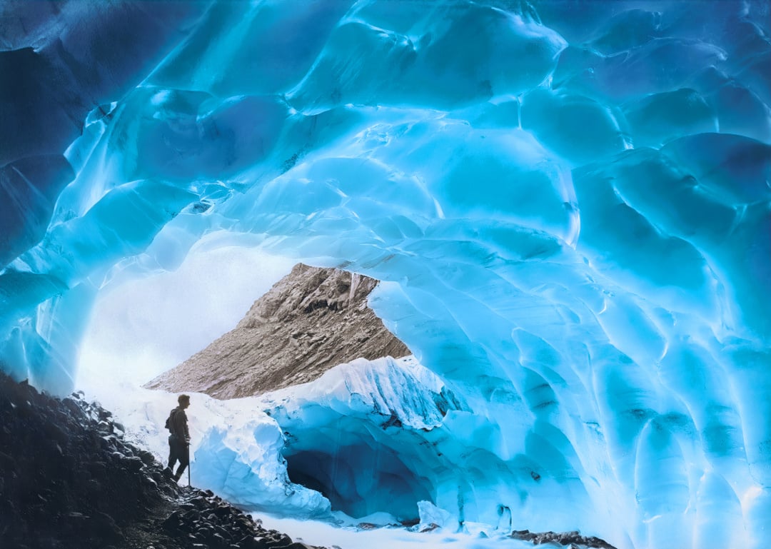 A person stands at the entrance of a cave made of shimmering blue ice, with a rocky mountain peak visible through the opening, creating a dramatic contrast of natural elements.