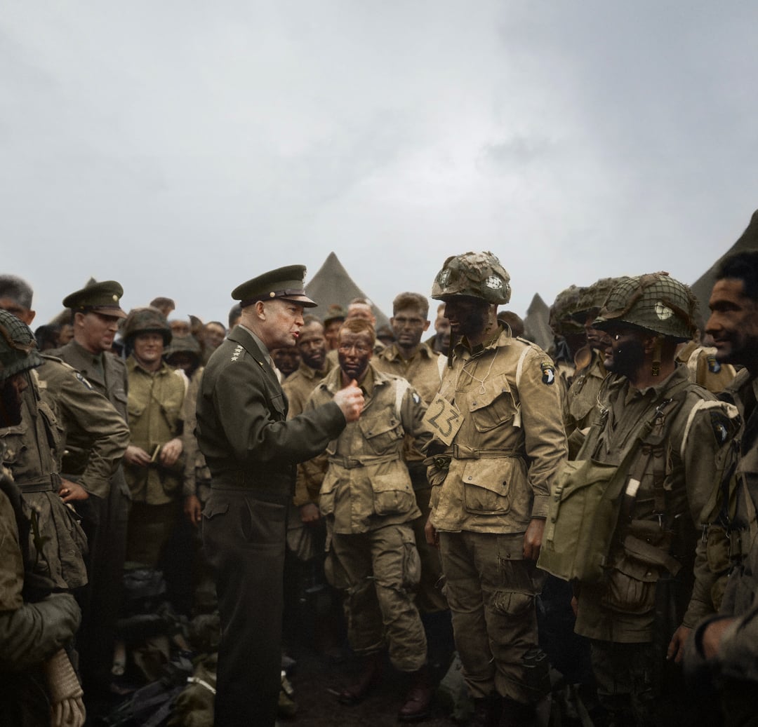 A military officer addresses a group of World War II soldiers in uniform, some with helmets and face paint, gathered closely outdoors under a cloudy sky. Tents are visible in the background.