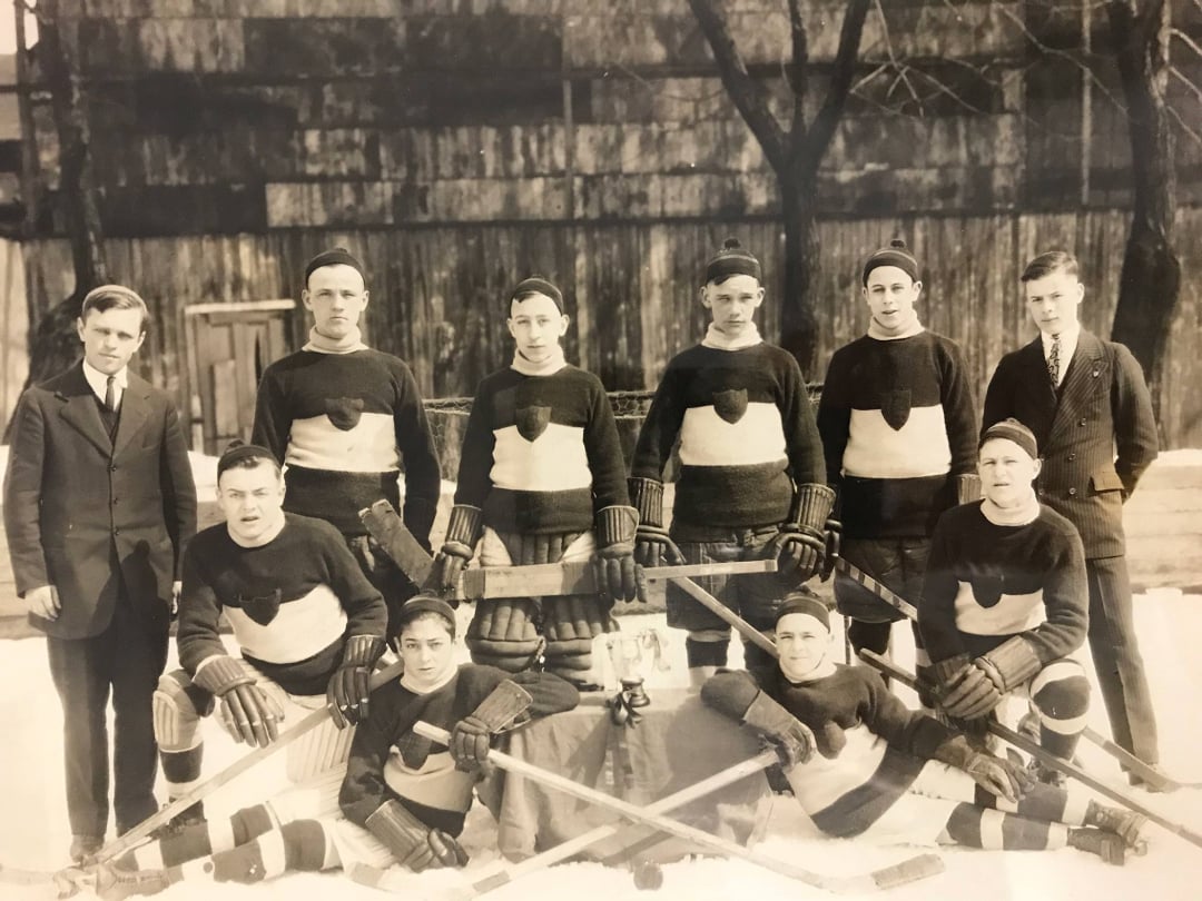 A vintage black-and-white photo of a men’s ice hockey team posing outdoors in winter, wearing matching jerseys with a round emblem, with two men in suits standing at each end. Hockey sticks and gear are arranged in front.