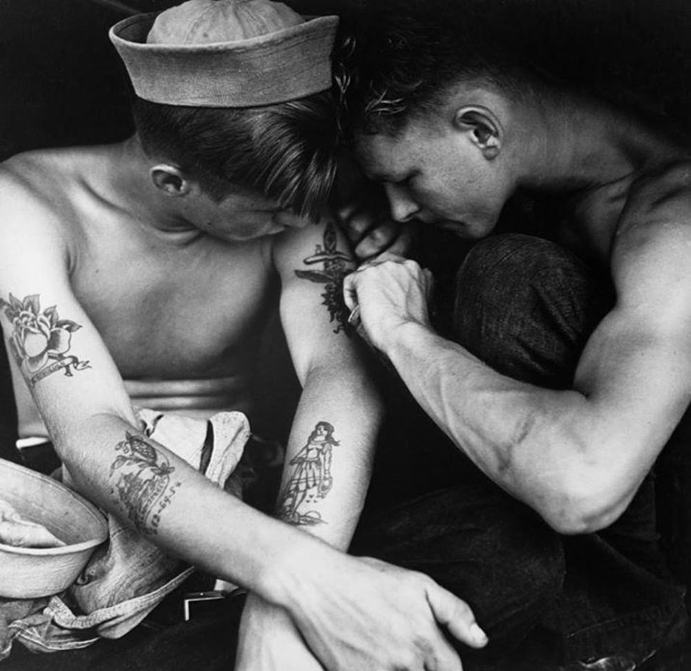 Two young men sit closely together, one shirtless and wearing a sailor hat, as the other tattoos his upper arm. The shirtless man has several visible tattoos on his arms. The scene is intimate and focused.