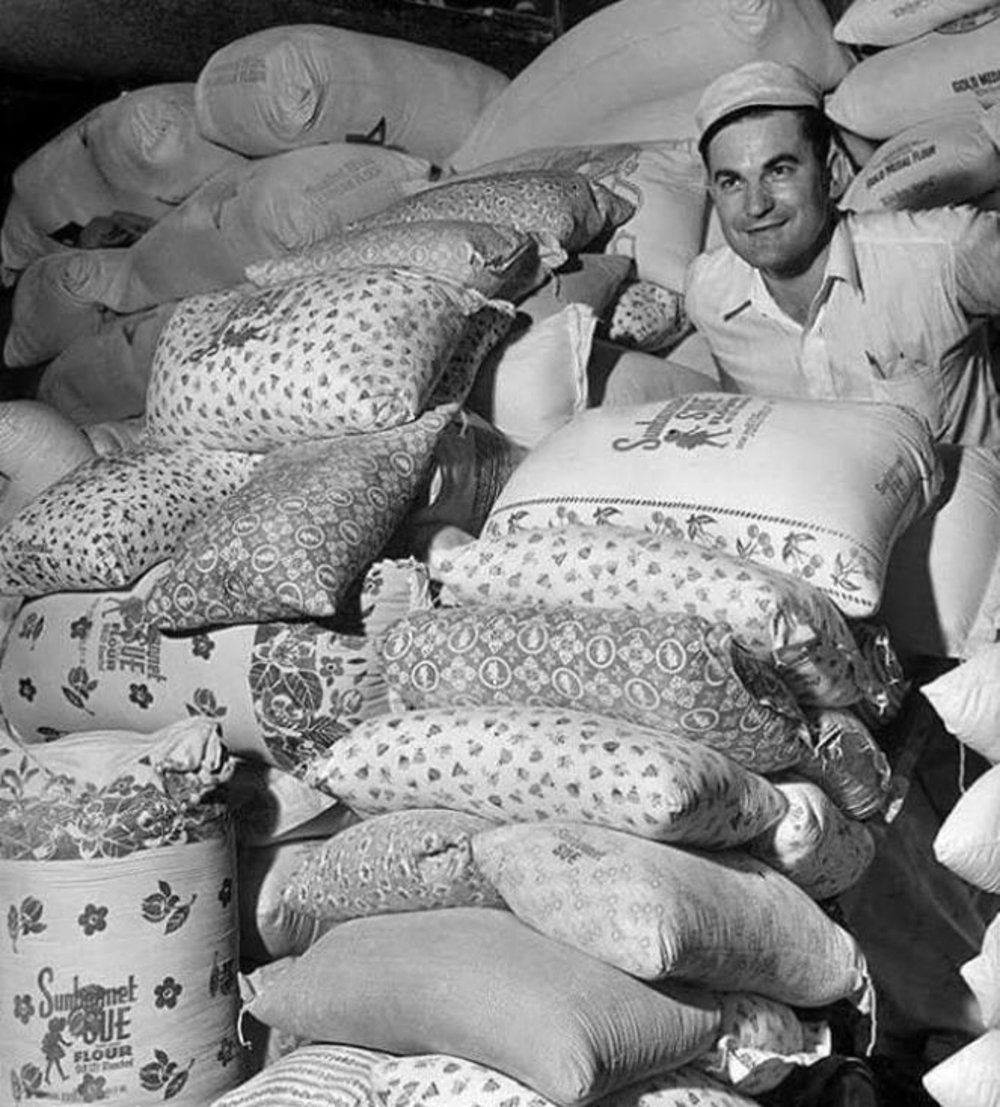 A man in a white shirt and cap stands behind a large pile of patterned flour sacks. The sacks have various floral and geometric designs, filling most of the scene. The setting appears to be a warehouse or storage area.