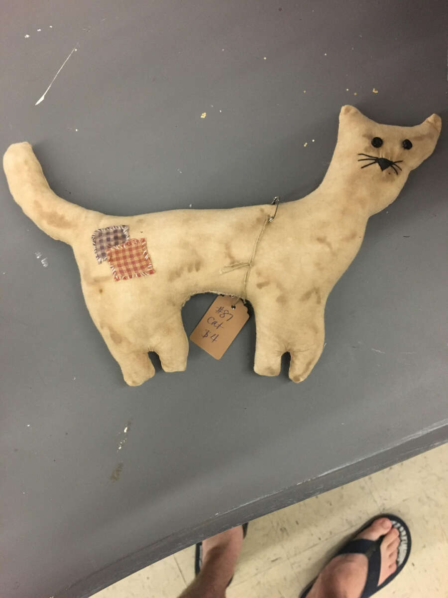 A handmade, tan fabric cat doll with button eyes, stitched whiskers, and two patchwork squares on its side lies on a gray surface. A tag reading “Cat #1” hangs from its belly. Someone’s feet in flip-flops are visible at the bottom.