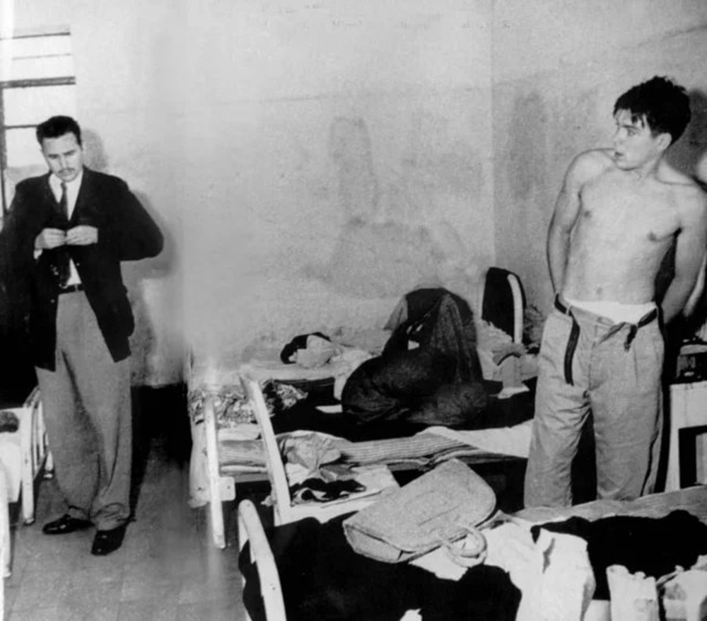 A black-and-white photo shows two men in a cluttered room with unmade beds. One man, dressed in a suit, stands on the left, while the other, shirtless, stands on the right, adjusting his pants.