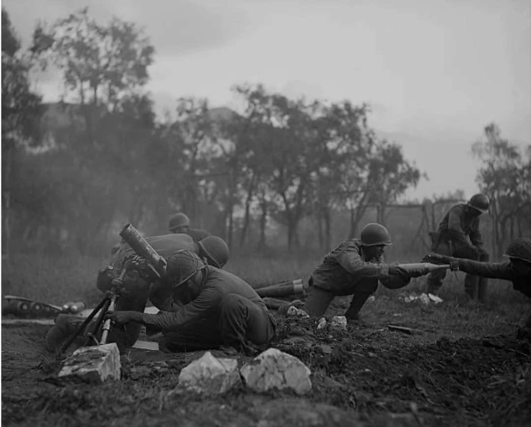 Five soldiers in helmets operate a mortar and prepare to fire while crouched on a dirt field, surrounded by rocks and trees, with smoke and haze in the background. The scene appears tense and wartime.