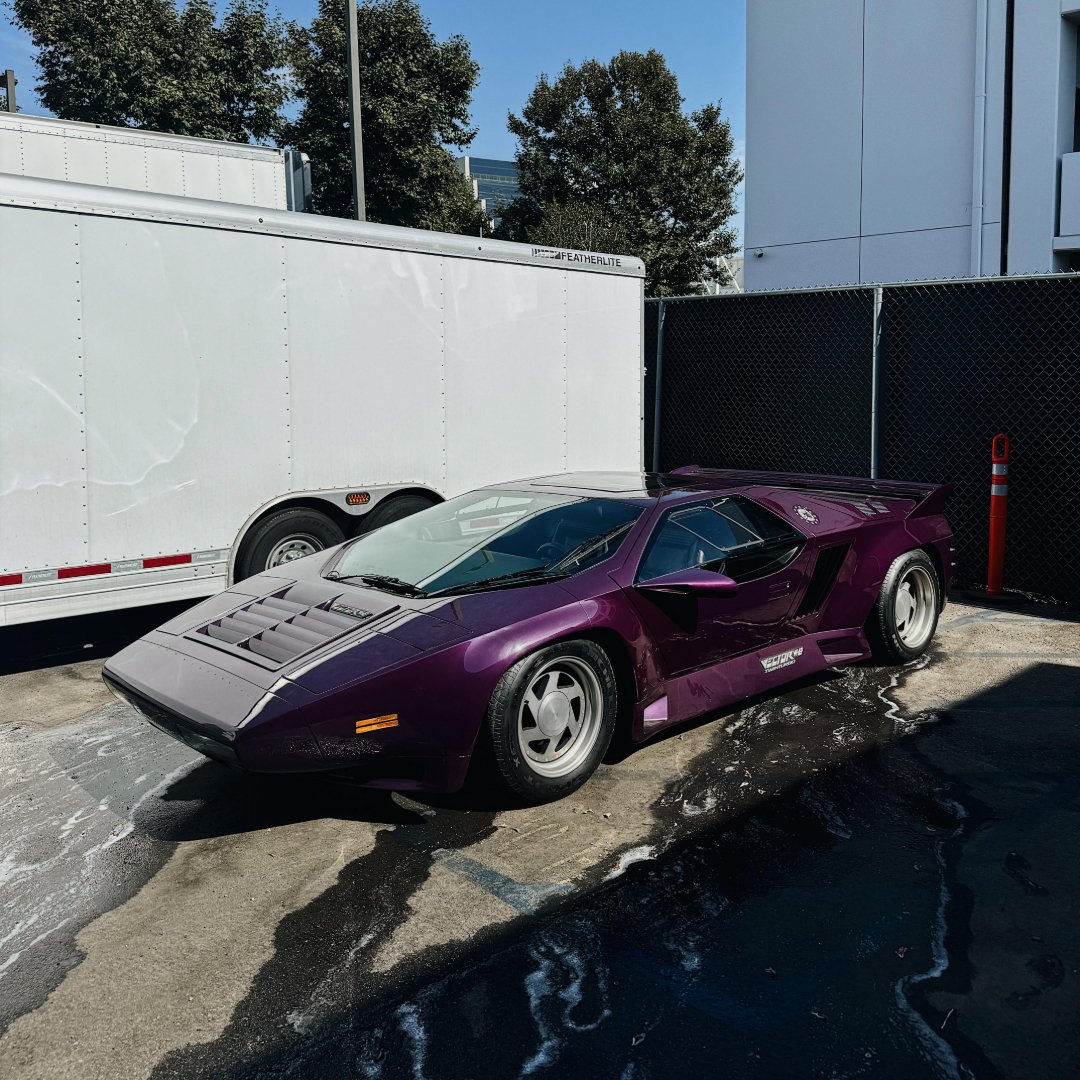 A purple sports car with sharp, angular lines is parked next to a white trailer in a sunlit lot. Trees and buildings are visible in the background.