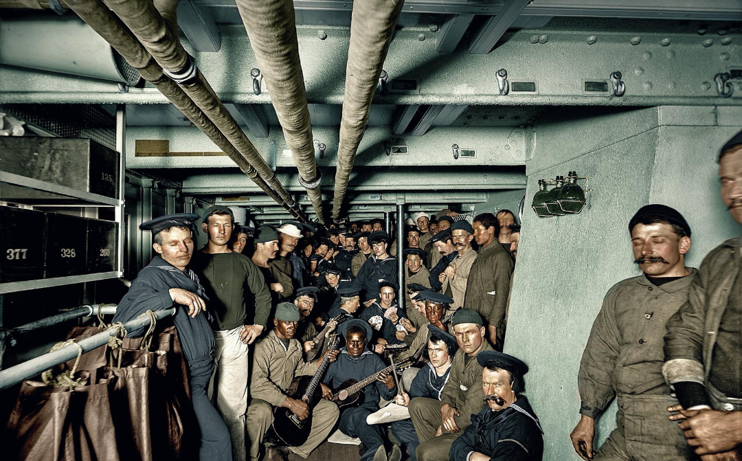 A group of sailors in uniform relax below deck, some sitting and some standing. Several play musical instruments, including a guitar and accordion, while others watch or chat under overhead pipes in the confined space.
