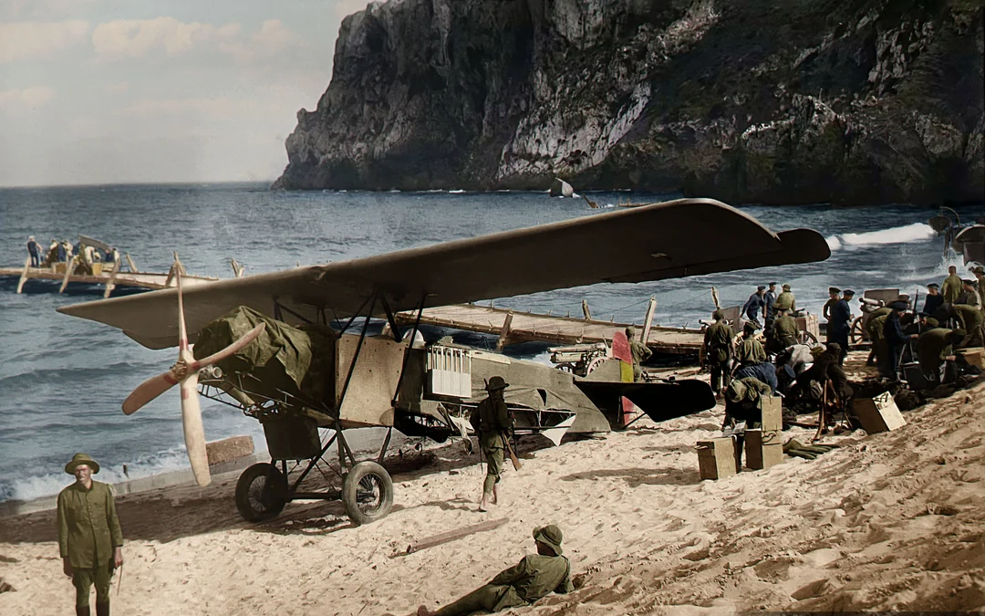 A vintage biplane sits on a sandy beach near the ocean, surrounded by uniformed soldiers and equipment. Rocky cliffs rise in the background, and wooden boats are visible along the shoreline.