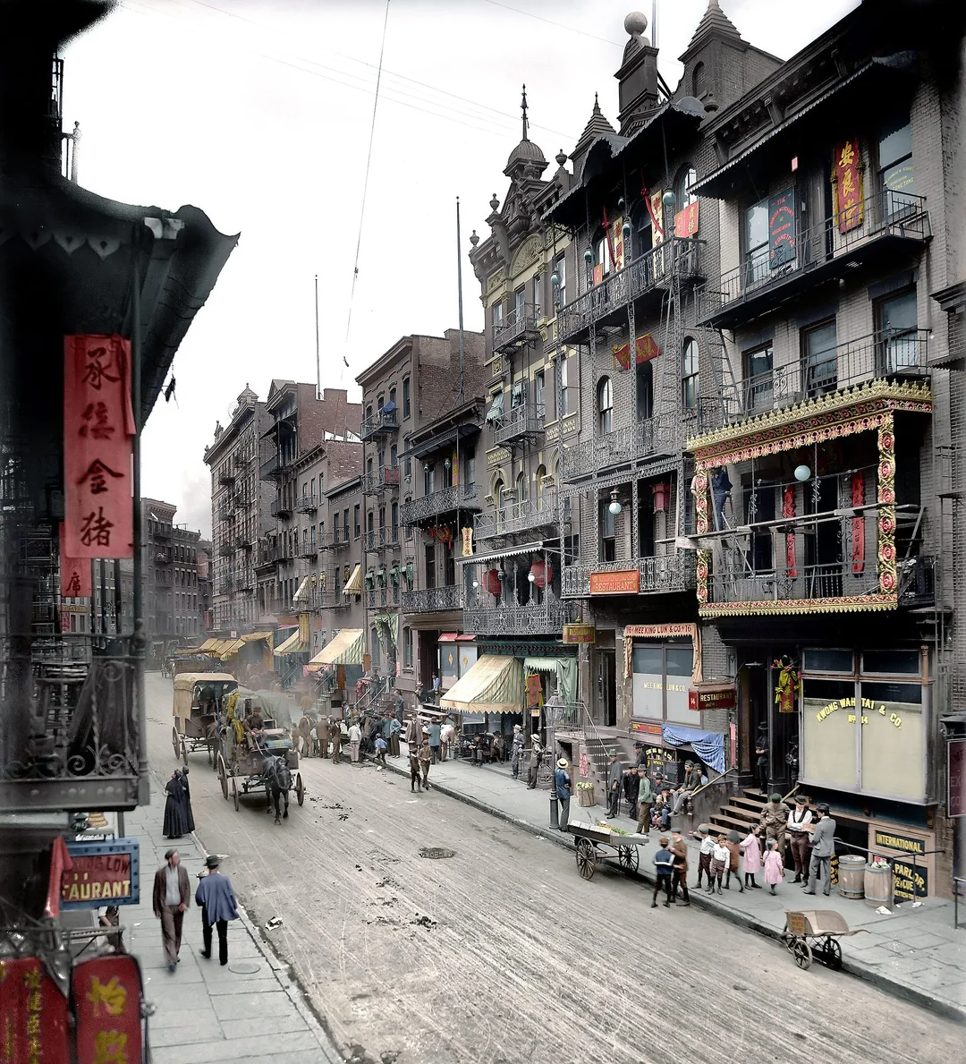 A bustling street scene in early 20th-century Chinatown, with ornate buildings, horse-drawn carriages, shop signs in Chinese, pedestrians, and groups of people gathered along the sidewalk.