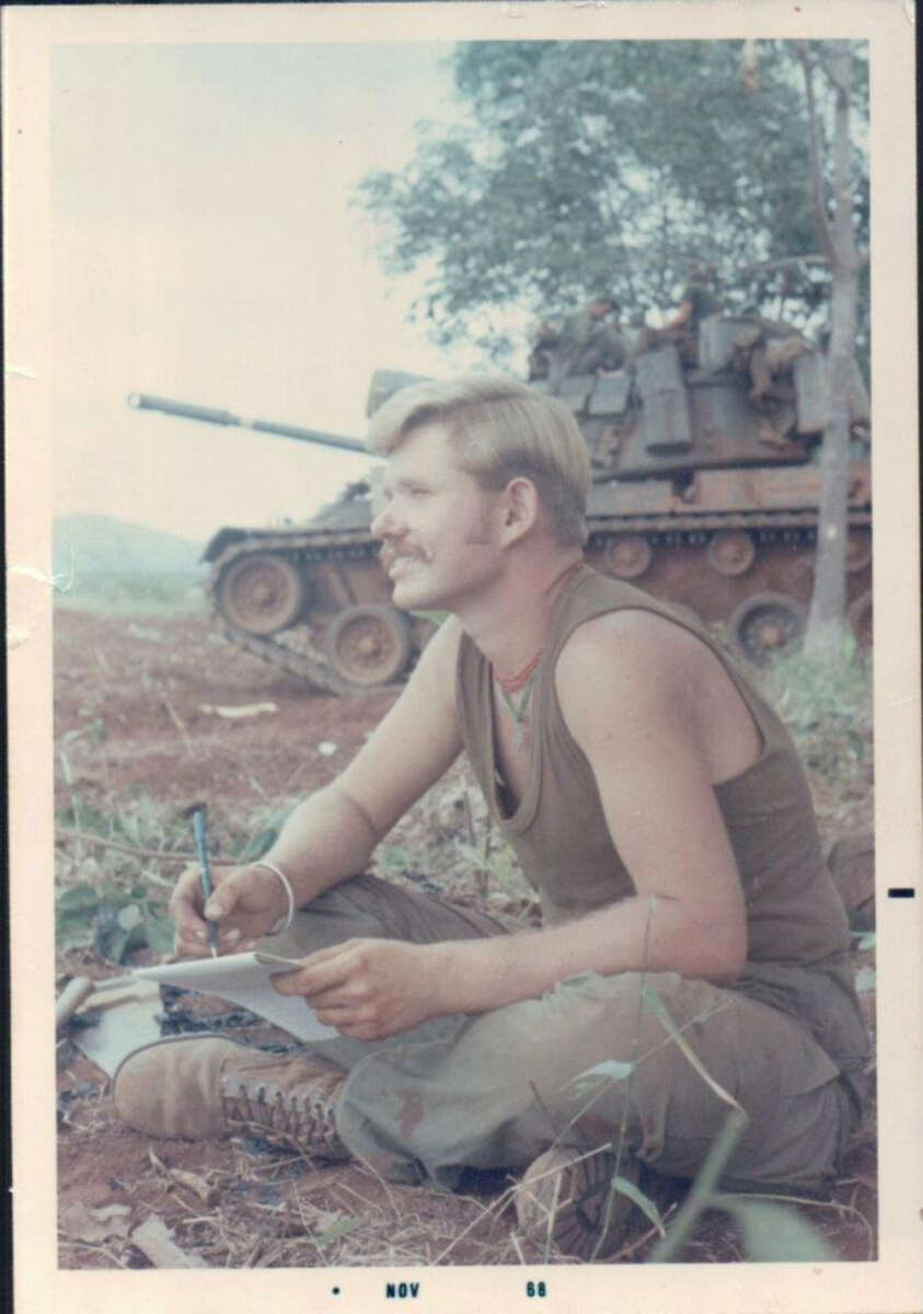 A man in a sleeveless shirt and military pants sits on the ground writing, with a tank and several soldiers in the background under a tree. The photo appears to be from the 1970s or 1980s.