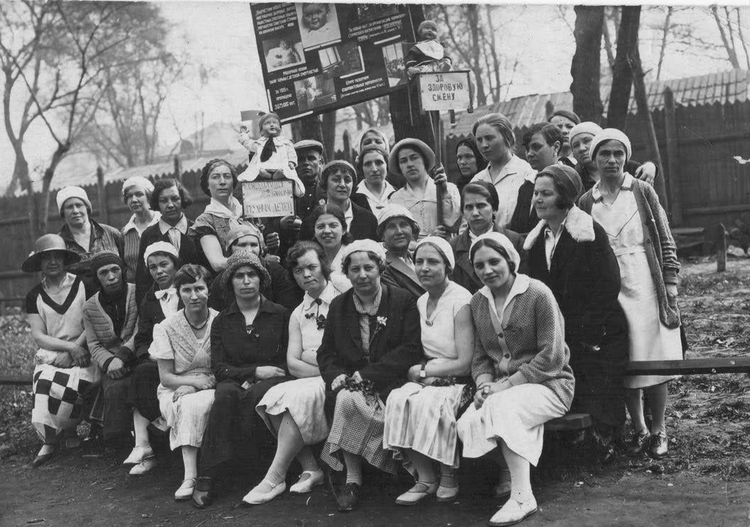 A group of women in 1920s-style clothing and headscarves pose outdoors, seated and standing, with trees and wooden fence in the background. Some hold posters with Cyrillic writing and a baby doll on top.