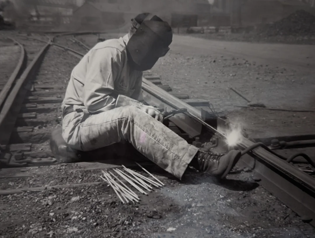 A person wearing protective gear welds a section of railroad track while sitting on the ground. Several welding rods are spread beside them, and sparks are visible from the welding torch.