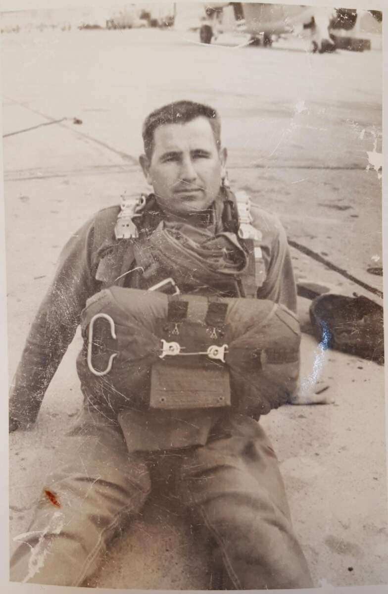 A man in a military uniform and parachute gear sits on the ground of an airfield, looking at the camera. The image is in black and white and appears to be an old photograph.