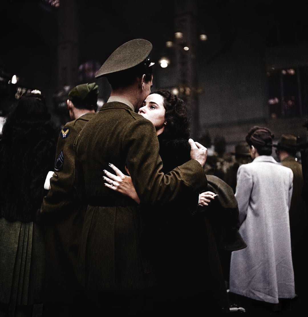 A woman embraces a soldier in uniform at a crowded train station, gazing up at him. Other people, some in coats and military uniforms, stand nearby, creating a poignant scene of farewell or reunion.