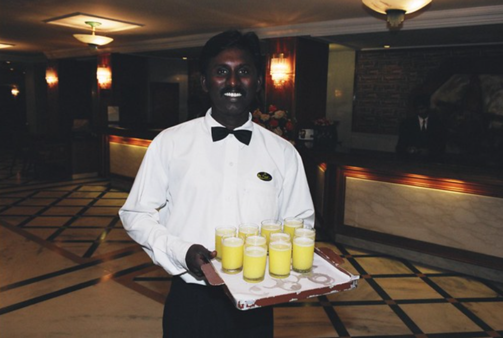 A smiling waiter in a white shirt and black bow tie holds a tray with several glasses of yellow drink, standing in a well-lit hotel lobby with marble floors and a reception desk in the background.