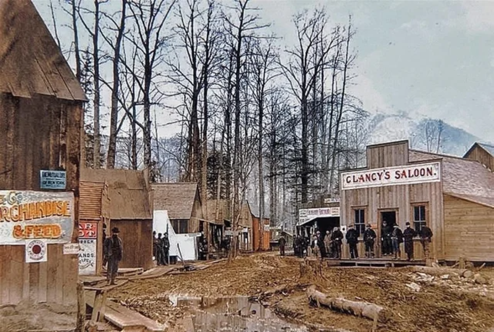 A muddy street in a small frontier town, lined with wooden buildings and leafless trees. People gather outside Clancy's Saloon; signs for various businesses are visible. Snowy mountains are seen in the background.