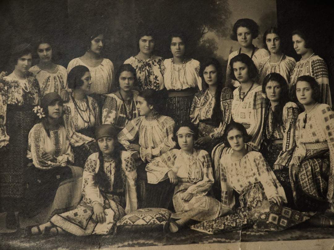 A vintage sepia photo of a group of women in traditional embroidered blouses and skirts, posing together indoors. Some are seated on the floor with patterned cushions while others stand behind.