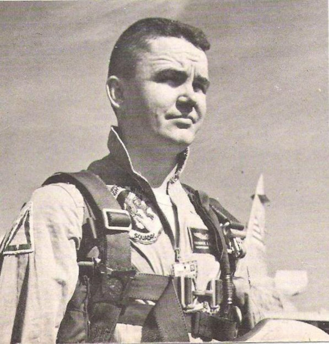 Black and white photo of a man in a flight suit and harness, standing outdoors. He looks slightly upward with a serious expression. A jet or missile is visible in the blurred background.