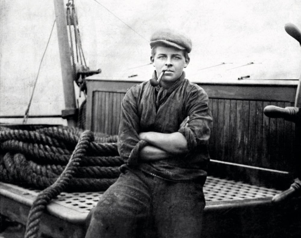 A young man wearing a cap and smoking a pipe sits with arms crossed on a wooden boat deck, with coiled ropes and nautical equipment visible in the background. The photo is black and white.