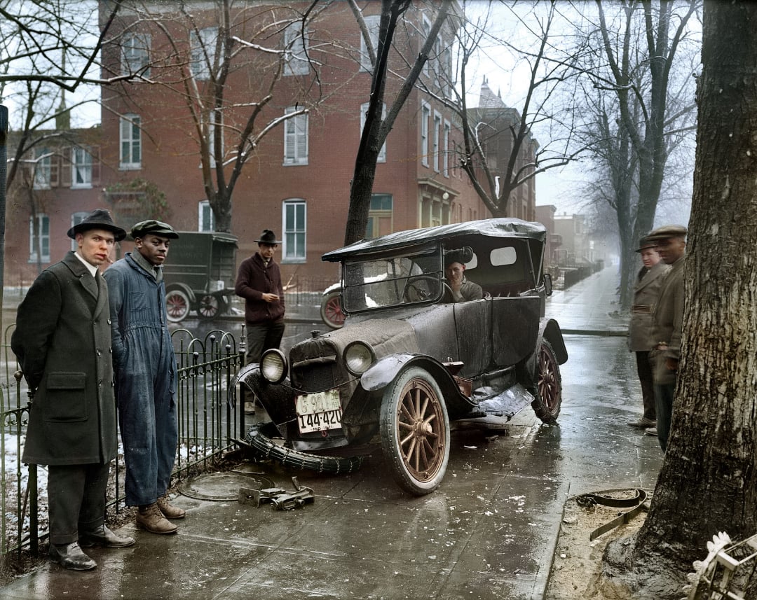 A vintage black car with front-end damage is parked on a wet city street. Four men stand nearby, two in suits and hats and two in work clothes. The scene is set in front of brick buildings on a rainy day.