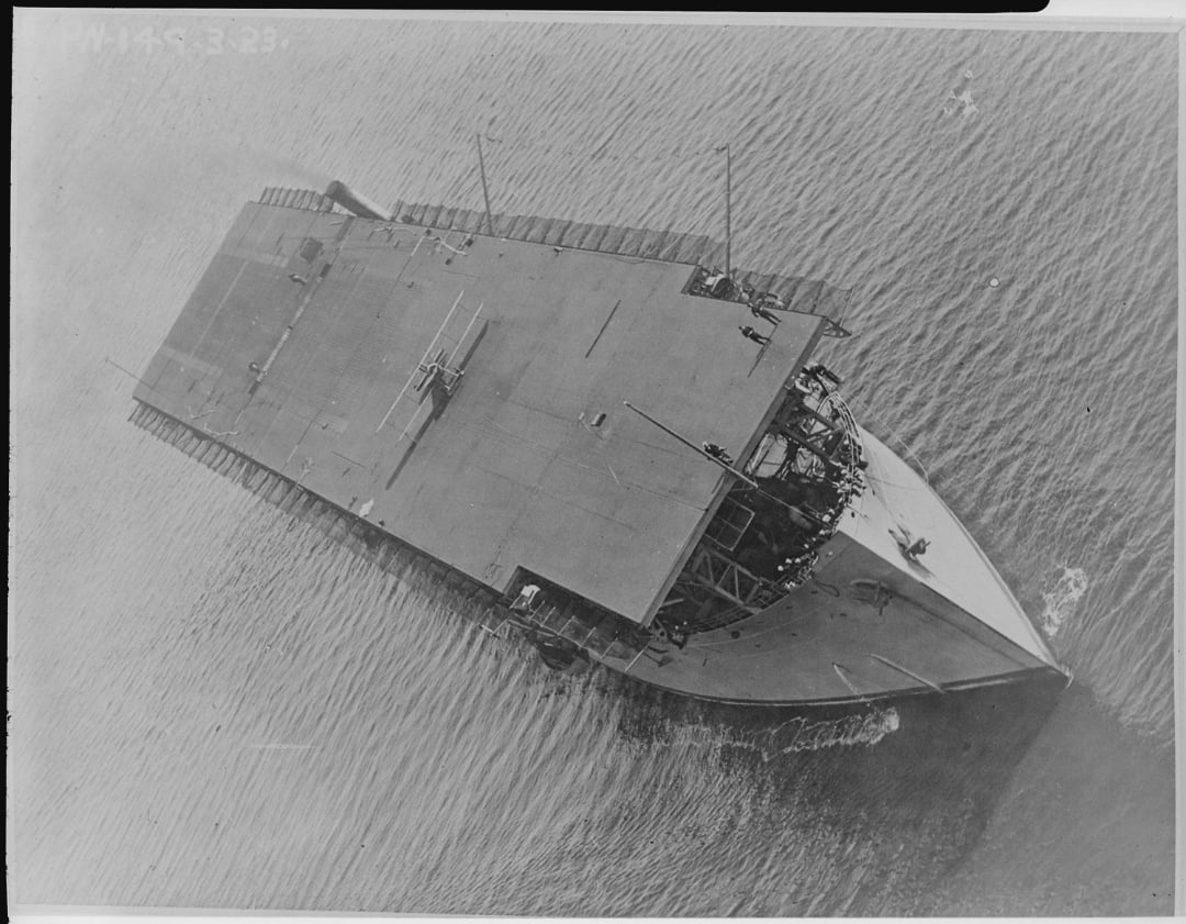 Aerial view of an early aircraft carrier sailing on the water, with a flat flight deck and several crew members visible on the deck. The ship’s bow is pointed toward the bottom right corner of the image.