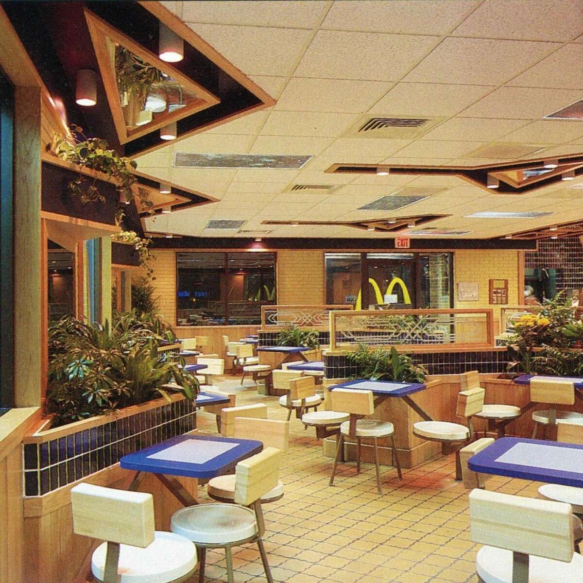 A retro fast food restaurant interior with blue and white tables, white stools, lots of greenery, wood paneling, and geometric ceiling lights. Large windows and a McDonald's logo are visible in the background.