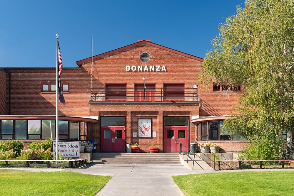 A brick school building with "BONANZA" written on top, two red doors, an American flag, and a sign reading "Bonanza School Grades K-12" in front. Green shrubs and a tree frame the entrance on a sunny day.