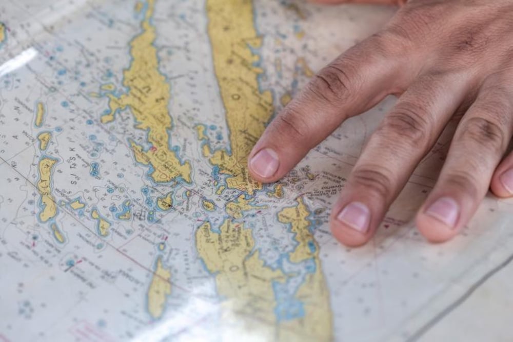 A close-up of a hand pointing to a specific location on a detailed nautical map with yellow landmasses and blue waterways.