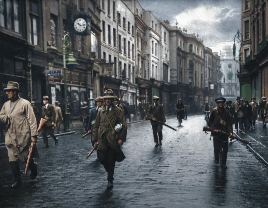 A group of men in coats and hats, some carrying rifles, walk along a wet, mostly empty city street lined with old buildings and shops under a cloudy sky. A clock is visible on a building. The image appears historical.