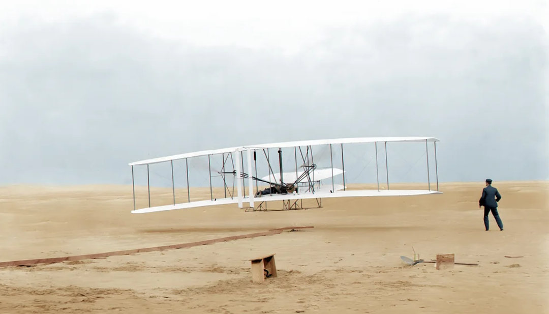 Historic black-and-white photo of the Wright brothers’ first powered flight in 1903, showing the Wright Flyer lifting off sandy ground with one person observing nearby.