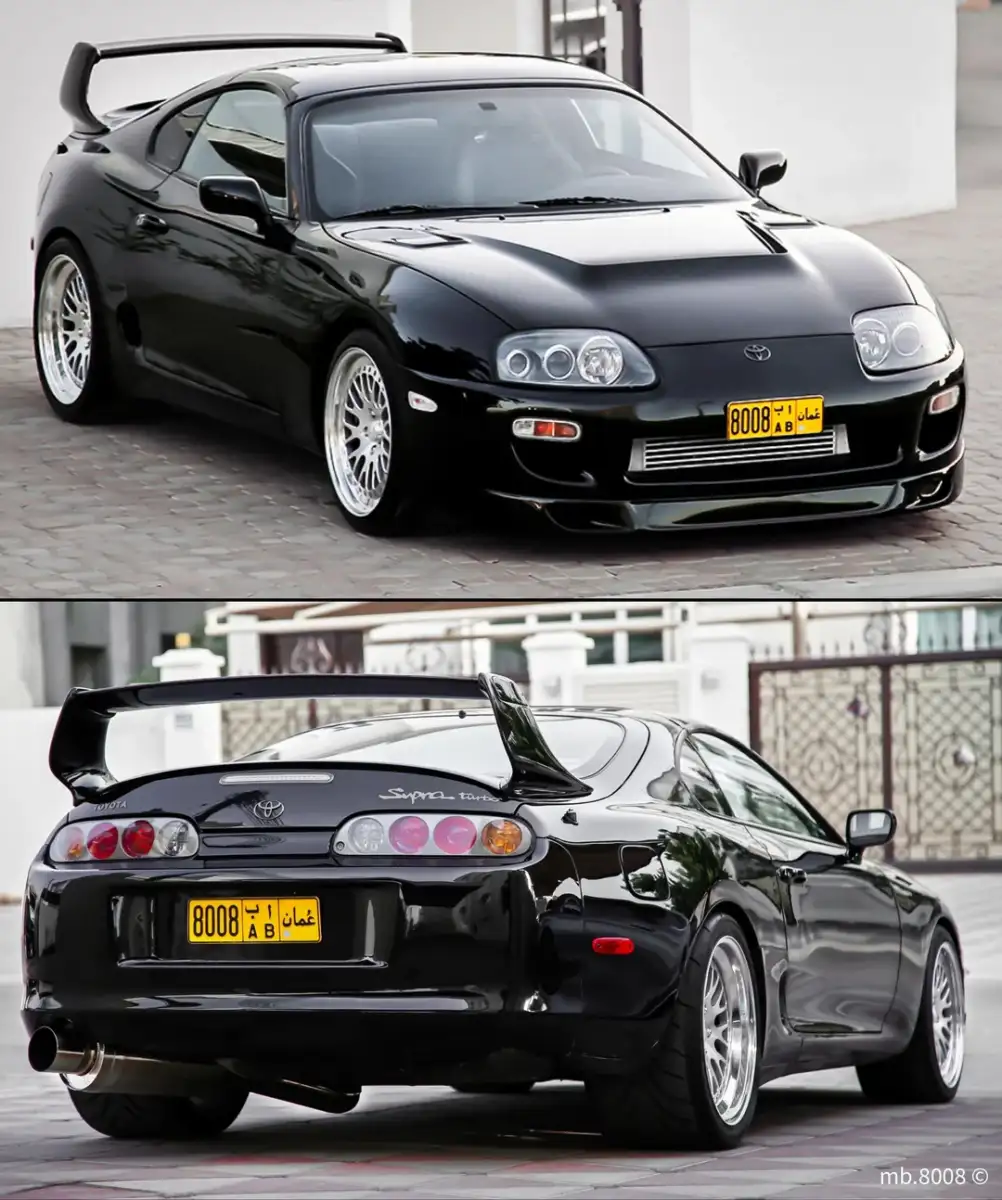 A black Toyota Supra sports car with a rear spoiler and yellow license plate is shown in two views: front-side and rear-side, parked on a paved surface. The car has shiny alloy wheels and a sleek, aerodynamic design.