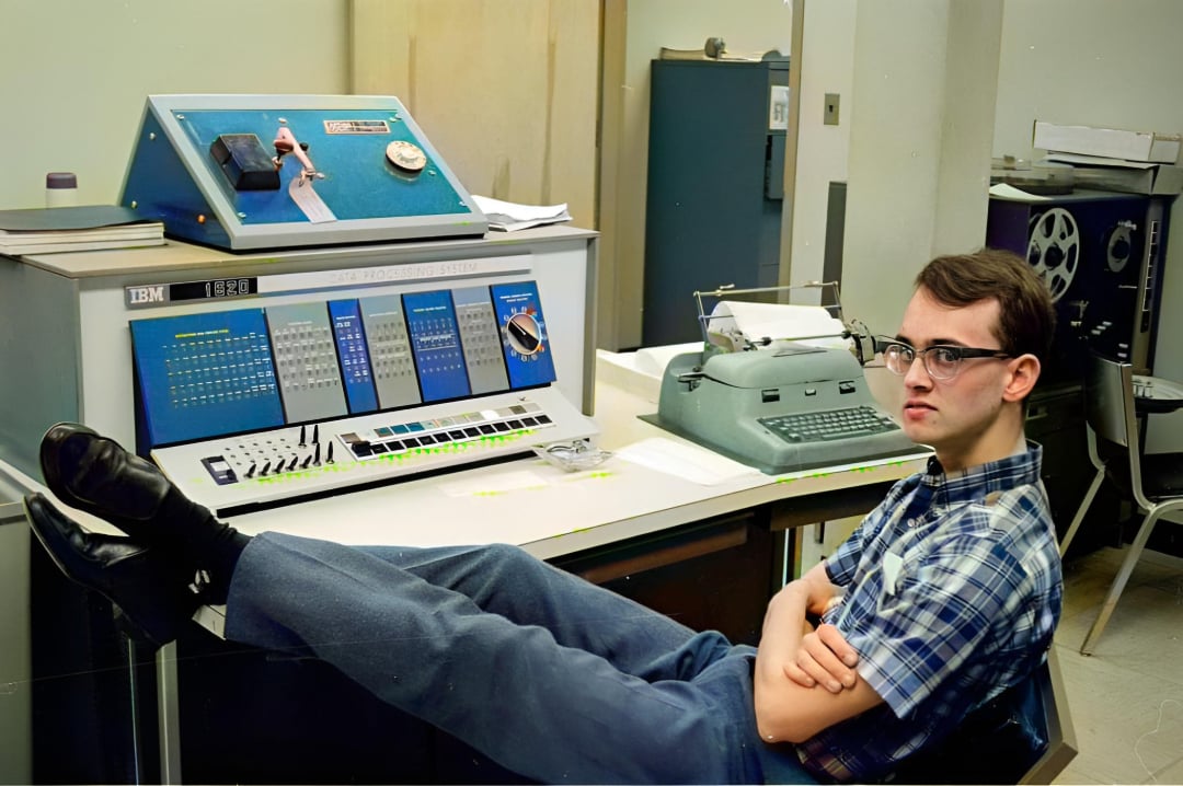 A young man with glasses, wearing a plaid shirt, sits with his legs up on a desk beside a vintage IBM computer console and an old typewriter in a retro office setting.