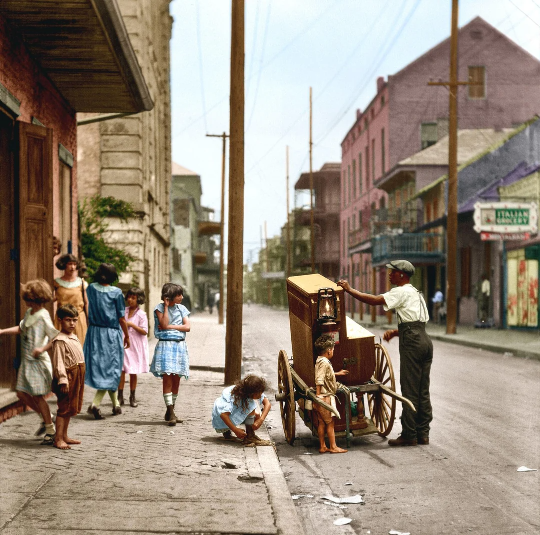 A group of children gather around an organ grinder’s cart on a city sidewalk, with colorful buildings and utility poles lining the street in the background.