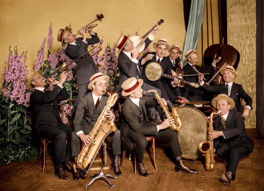 A vintage jazz band with eleven men in suits and straw boater hats play various instruments, including saxophones, trumpet, trombone, clarinet, upright bass, and drums, in front of a floral backdrop.