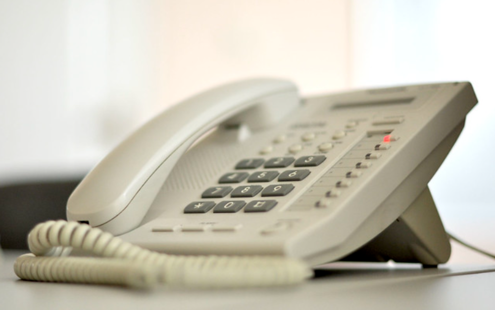 A close-up of a beige office landline telephone with large number buttons, a corded handset, and indicator lights, sitting on a white desk. The background is softly blurred.