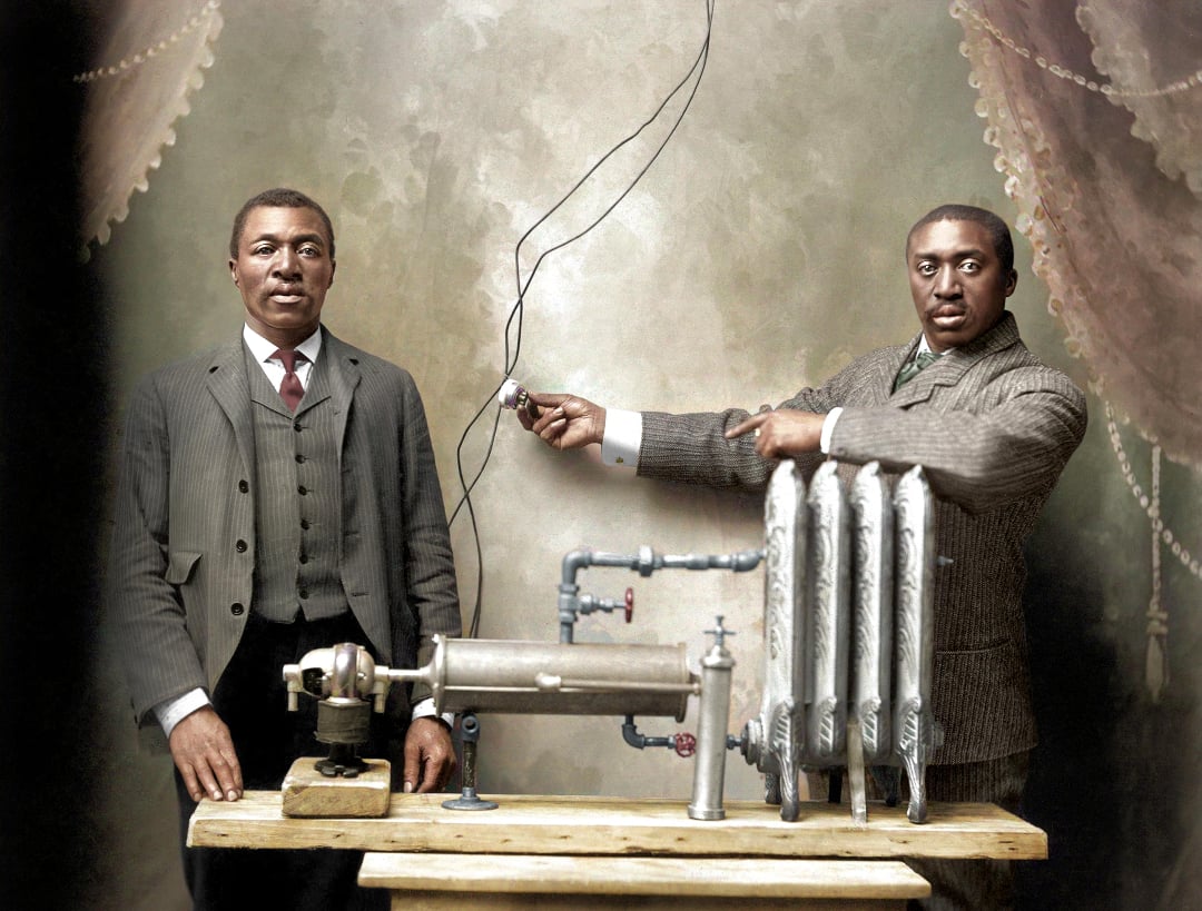 Two men in suits stand by an early heating system prototype with pipes, valves, and a radiator on a table; one man points at the device while the other stands beside, all set against a vintage painted backdrop.