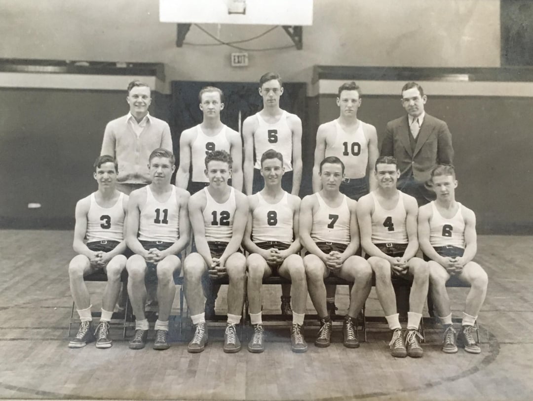Black-and-white photo of a vintage basketball team posing in a gym. Ten players in numbered uniforms sit and stand in two rows, with two men in suits standing at the ends. A basketball hoop is visible in the background.