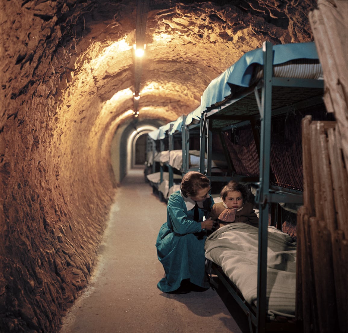 A woman in a blue dress comforts a child lying on a bunk bed in an underground tunnel lined with beds, illuminated by warm overhead lights.
