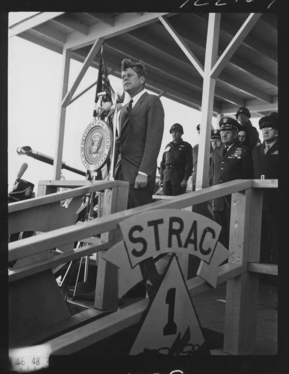 A man in a suit stands at a podium with the U.S. presidential seal, flanked by military officers on a stage. In the foreground, a sign reads "STRAC 1." Flags and microphones are visible.